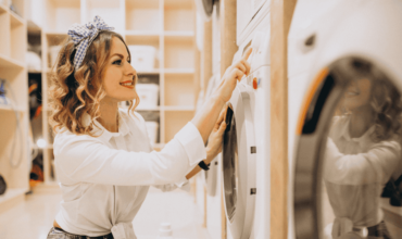 Happy woman with curly hair and a blue headband operating a commercial tumble dryer in a modern laundromat.