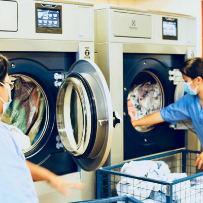 Staff members using commercial tumble dryers to handle laundry in a professional facility.