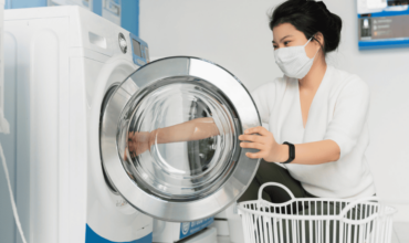 Woman in a white robe and protective mask loading laundry into an Electrolux Australia commercial washing machine, highlighting the appliance's use in a public laundry setting.