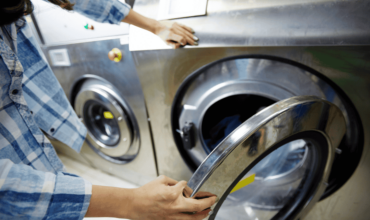 Close-up of a woman in a plaid shirt opening a commercial tumble dryer, using Electrolux Australia equipment in a busy laundromat setting.