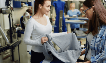 Two women in a textile factory inspecting a freshly ironed garment, with Electrolux Australia automatic ironing machines visible in the background, emphasizing efficiency in garment production.