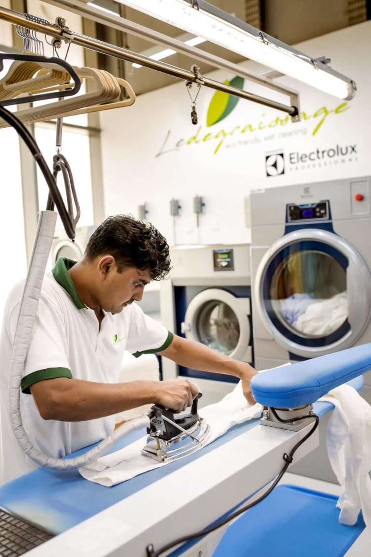 A man irons a white shirt in a well-lit laundry room, surrounded by cleaning supplies and washing machines.