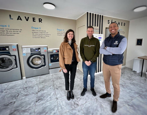 Three people, smiling, in the center of a new laundry shop, with washing machines and a dryer.